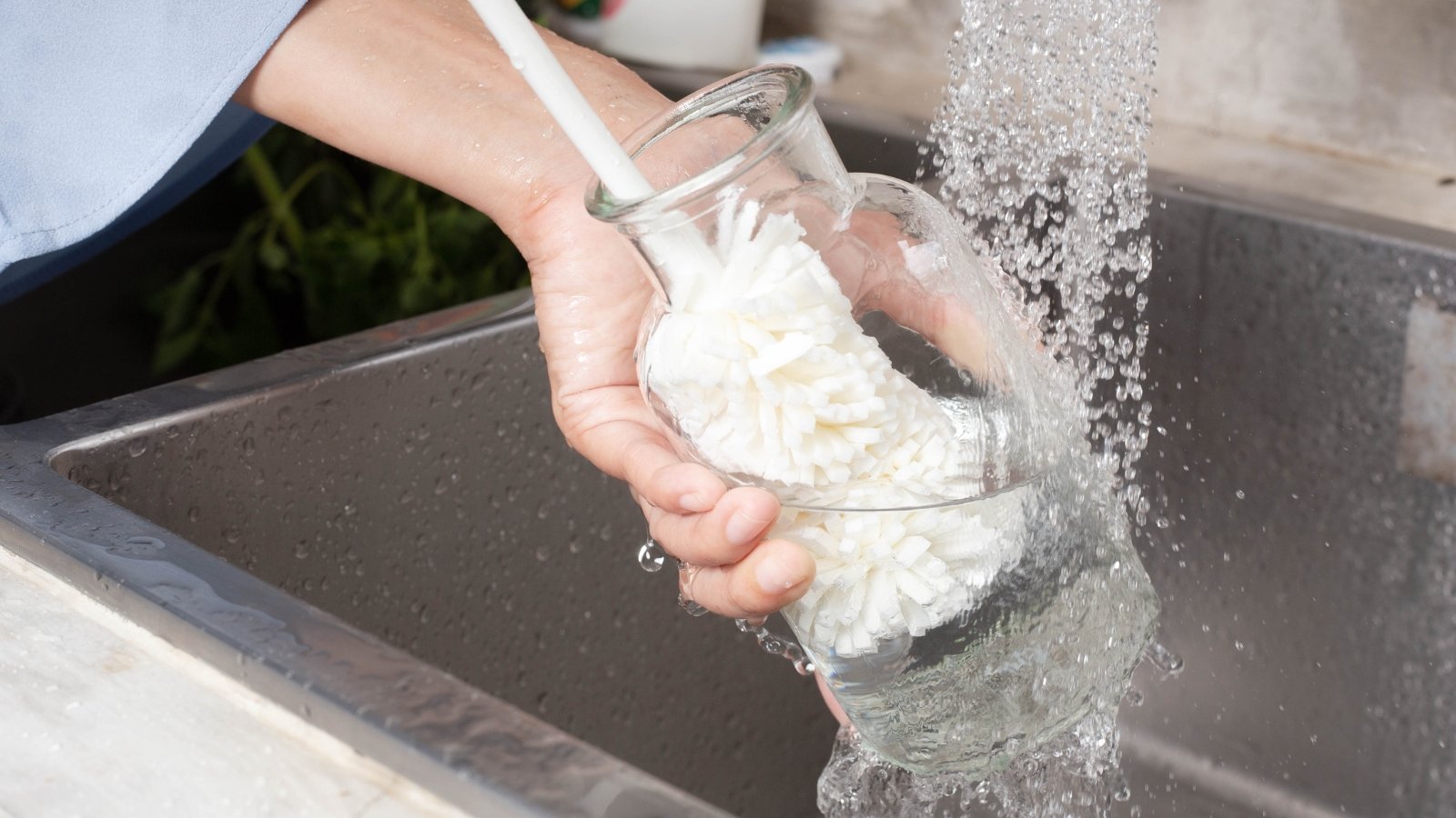 Hands rinsing a glass vase under running tap water, removing dirt.